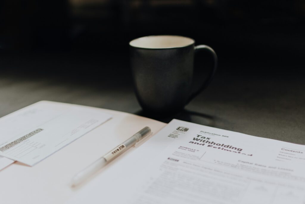 Close-up of a tax preparation scene featuring a pen, a tax withholding document, and an envelope laid out on a desk. A dark ceramic coffee mug sits in the background, creating a calm and focused workspace atmosphere.