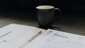 Close-up of a tax preparation scene featuring a pen, a tax withholding document, and an envelope laid out on a desk. A dark ceramic coffee mug sits in the background, creating a calm and focused workspace atmosphere.