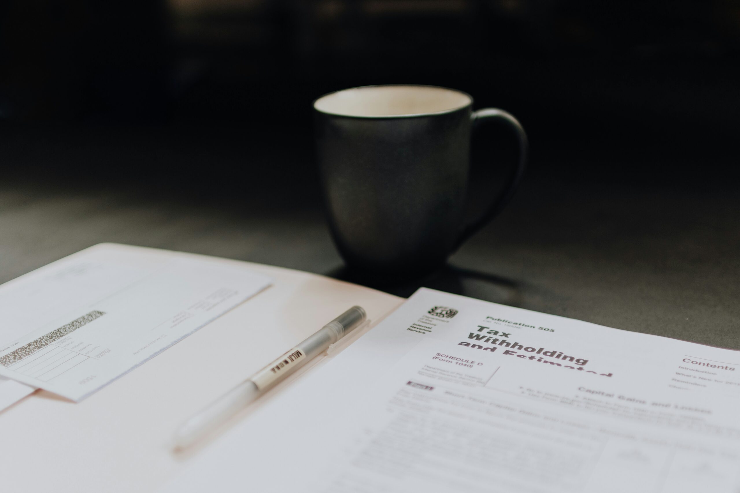 Close-up of a tax preparation scene featuring a pen, a tax withholding document, and an envelope laid out on a desk. A dark ceramic coffee mug sits in the background, creating a calm and focused workspace atmosphere.