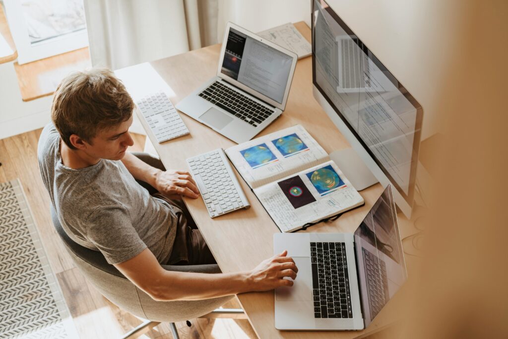 A man works at a modern desk setup with three computers and a notebook filled with colorful data visualizations. This workspace is organized for multitasking and data analysis