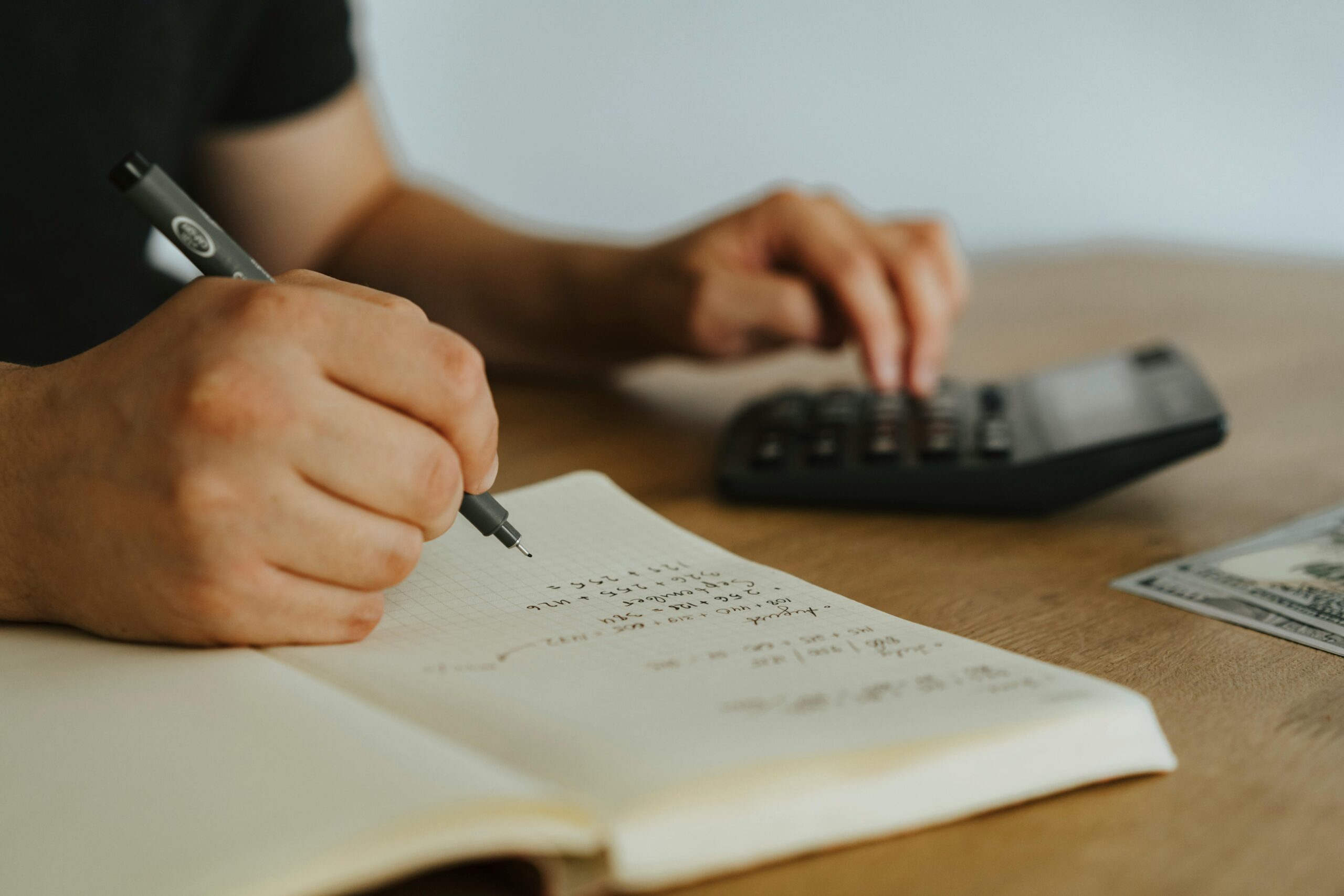 Close-up of a person writing calculations in a notebook with one hand while using a calculator with the other. A U.S. hundred-dollar bill lies nearby on the wooden table, suggesting a financial or budgeting task in progress.