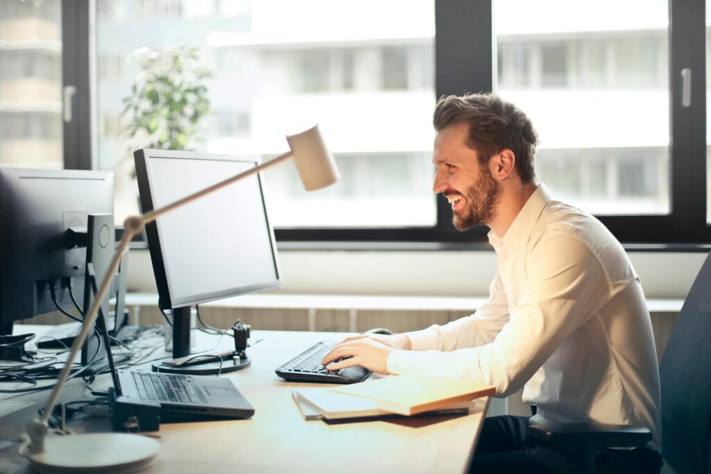Cheerful man in a white dress shirt working at his desk with multiple monitors in a bright, modern office. Natural light pours in through large windows, creating an energizing and productive workspace