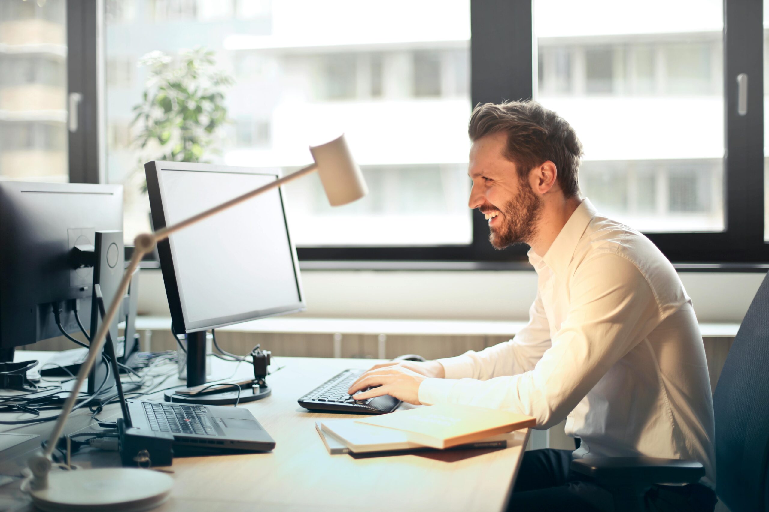 Cheerful man in a white dress shirt working at his desk with multiple monitors in a bright, modern office. Natural light pours in through large windows, creating an energizing and productive workspace
