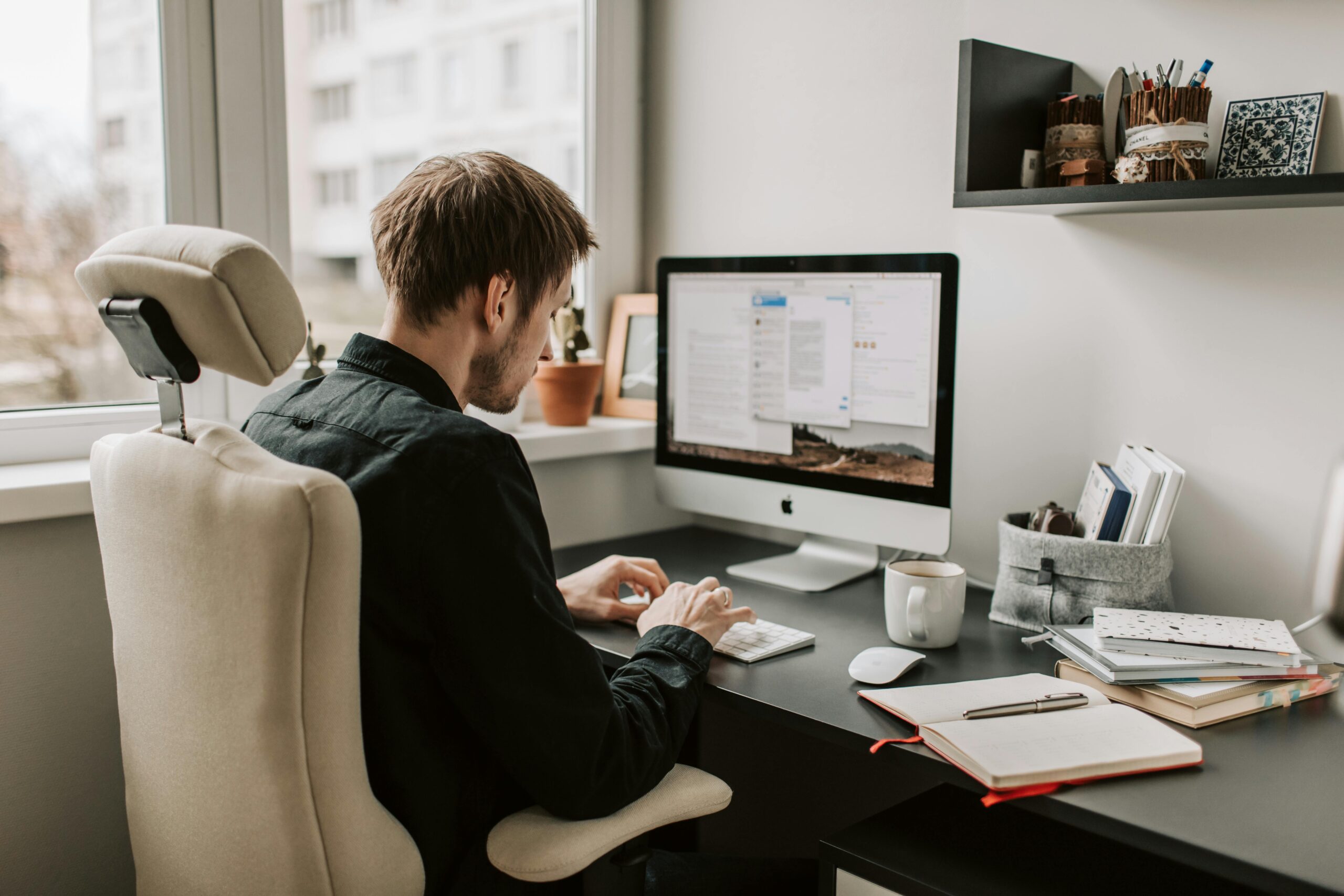 A focused man sits in a cozy home office, working on a desktop computer. The desk is organized with notebooks, a coffee mug, and small storage containers, creating a productive and calm environment. Natural light filters through the window beside him.
