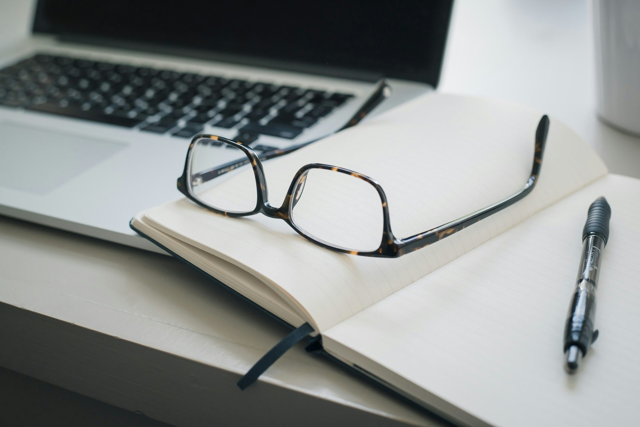 Close-up of a workspace featuring a pair of tortoiseshell eyeglasses resting on an open lined notebook. A black pen lies beside the notebook, and a laptop is partially visible in the background.